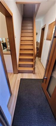 A bright hallway with a wooden staircase in the background. The floor is made of parquet and there is a doormat at the entrance.