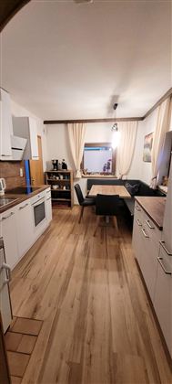 A modern kitchen with a light wooden floor and white furniture. A dining area with a table and a bench is also visible.
