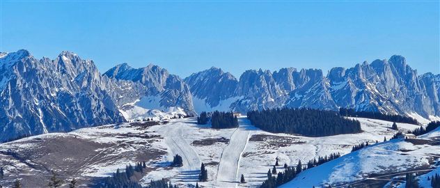 A snow-covered mountain landscape under a clear blue sky. In the foreground, ski slopes and coniferous trees can be seen.
