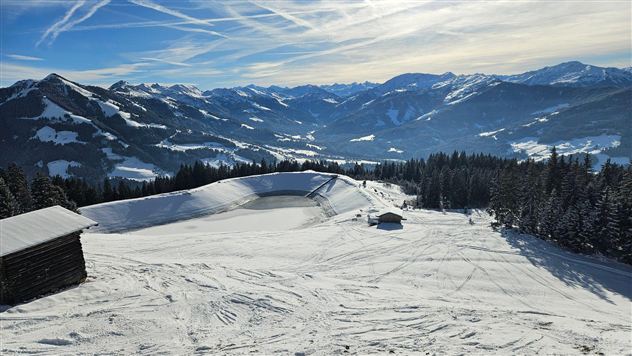 A snow-covered mountain with wide, green valleys and a clear, blue sky view. In the foreground, a few wooden cabins are visible.