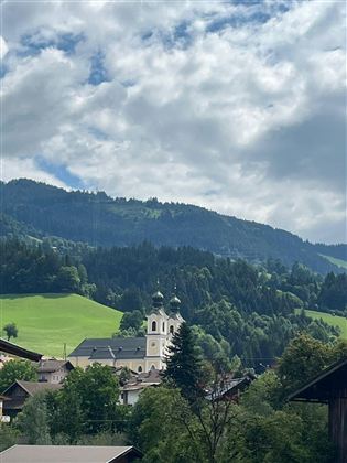 A picturesque landscape with gentle hills and a small church in the foreground. In the background, wooded mountains and a cloudy sky can be seen.
