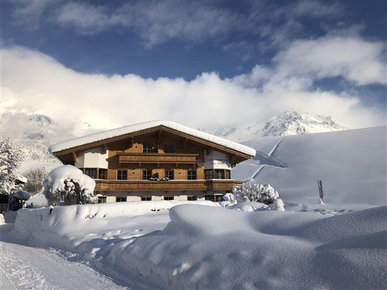 A cozy wooden house in the snow, surrounded by a wintry landscape. The mountains are visible in the background, and the sky is partly cloudy.