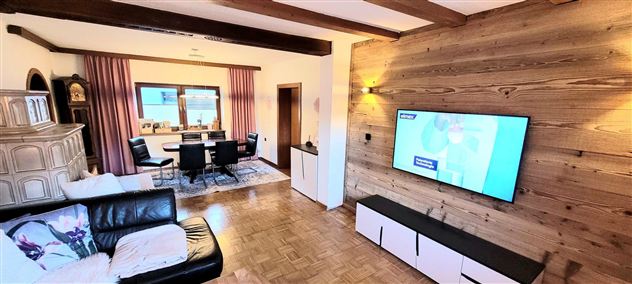 A cozy living room with a large couch and a modern television. The room is bright and has wooden beams as well as an elegant dining area.