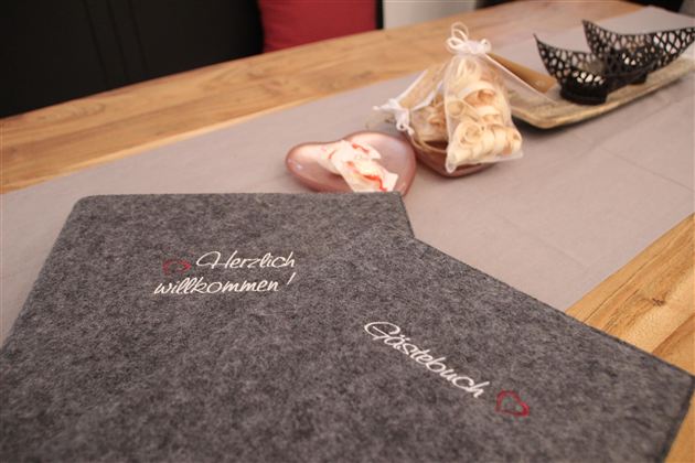 Ein schön gedeckter Tisch mit einem grauen Menübuch und einer herzlichen Willkommensbotschaft. Im Hintergrund sind gefüllte Tüten und dekorative Elemente angeordnet.