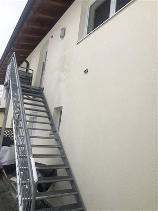 Ein grauer, metallener Treppenaufgang führt zu einem Eingang an einer hellen Wand. Über der Treppe befinden sich zwei Wandleuchten.
