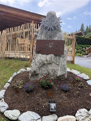 A large rock with a metal plaque displaying the name "Auen." Colorful flowers and stones decorate the area around the rock.