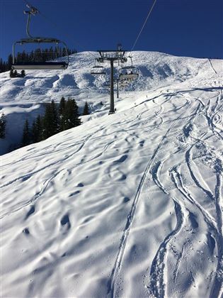 Eine schneebedeckte Piste mit Skispuren und einem Skilift im Hintergrund. Der klare blaue Himmel ergänzt die winterliche Landschaft.