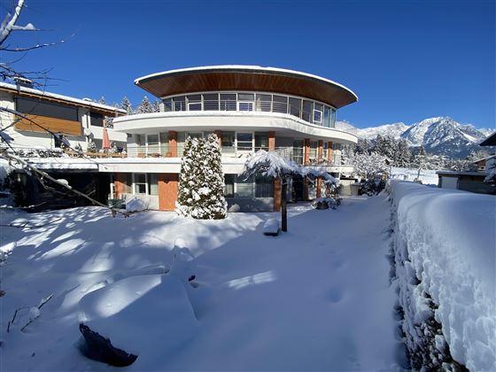 Ein modernes Haus in einer schneebedeckten Landschaft. Die Umgebung ist von Bergen und klarem blauen Himmel umgeben.