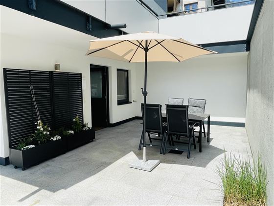 A modern terrace with a large sun umbrella and a dining area. In the background, plants and a simple wall can be seen.