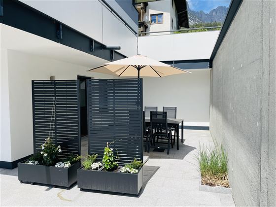 A modern terrace with a dining table and a sunshade.  
Surrounded by decorative plants and a well-kept wall.