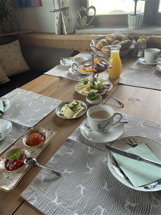 Ein schön gedeckter Frühstückstisch mit Tassen, Geschirr, Marmelade, Käse und frischem Obst. Im Hintergrund sind Brötchen und ein Glas Saft zu sehen.