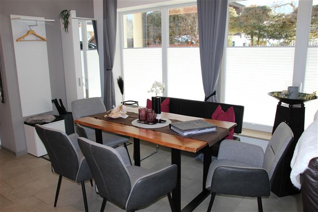 Ein moderner Essbereich mit einem stilvollen Holztisch, einer Bank und vier grauen Stühlen. Große Fenster sorgen für viel Tageslicht.