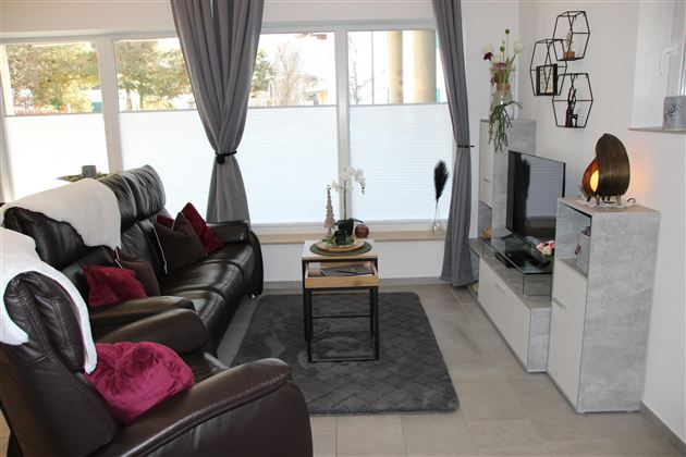 Ein modernes Wohnzimmer mit einem bequemen schwarzen Sofa und dekorativen Kissen. Der Raum hat große Fenster und einen Fernseher auf einem schlichten Möbelstück.