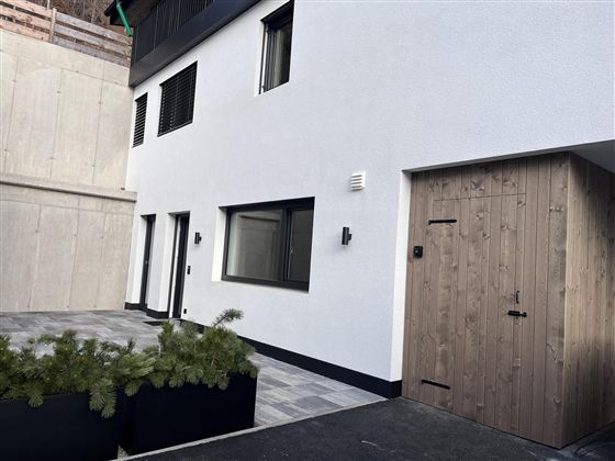 Ein modernes, weißes Gebäude mit großen Fenstern und einer Holzgarage. Der Bereich ist sauber und gut gestaltet, umgeben von Pflanzkübeln.