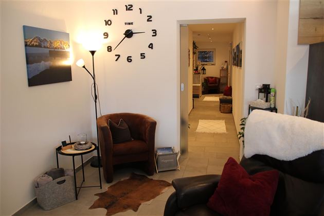 Ein einladendes Wohnzimmer mit einem bequemen Sessel, einem Tisch und einer großen Wanduhr. Der Raum hat einen hellen, freundlichen Eindruck und führt zu einem weiteren Bereich.