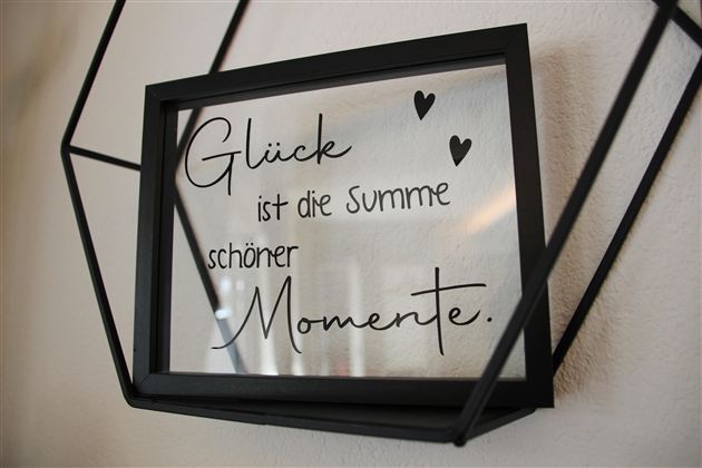 Ein gerahmtes Glasbild mit der Aufschrift: „Glück ist die Summe schöner Momente.“. Der Rahmen ist schwarz und hat eine moderne Form.