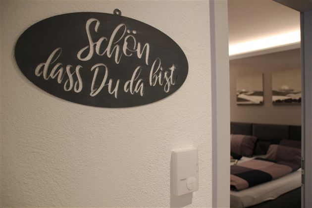 Ein Willkommensschild mit der Aufschrift „Schön, dass Du da bist“. Im Hintergrund ist ein gemütlicher Raum mit einem Bett und sanften Farben zu sehen.