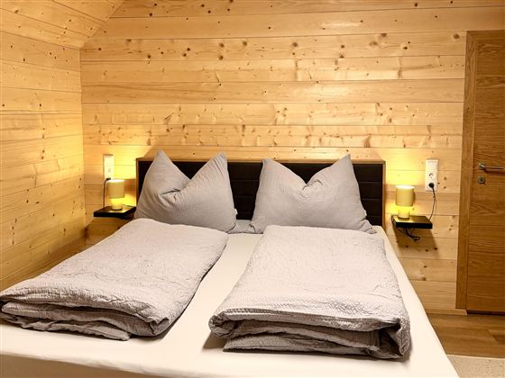 A cozy bedroom with wooden paneling and a large double bed. The bedding is in soft shades of gray, and there is gentle light from bedside lamps.