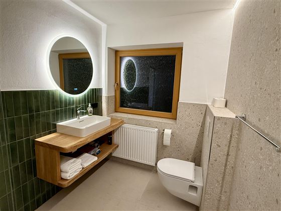 Ein modernes Badezimmer mit einem großen Spiegel und einem Fenster. Die Wände sind teilweise mit grünen Fliesen verkleidet, und es gibt Holzregale mit Handtüchern.