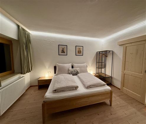 Ein modernes Schlafzimmer mit einem Doppelbett, zwei Kissen und einer neutralen Farbpalette. An der Wand hängen zwei Bilder, und ein Regal aus Metall steht im Raum.