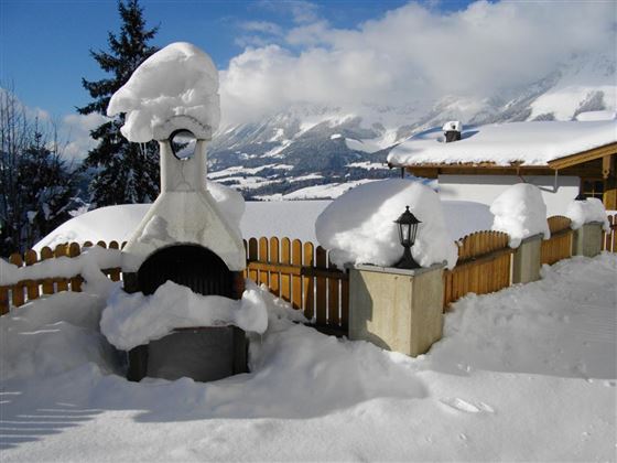 Eine verschneite Landschaft mit einem Grill und Laternen, die von Schnee bedeckt sind. Im Hintergrund sind Berge und ein blauer Himmel zu sehen.