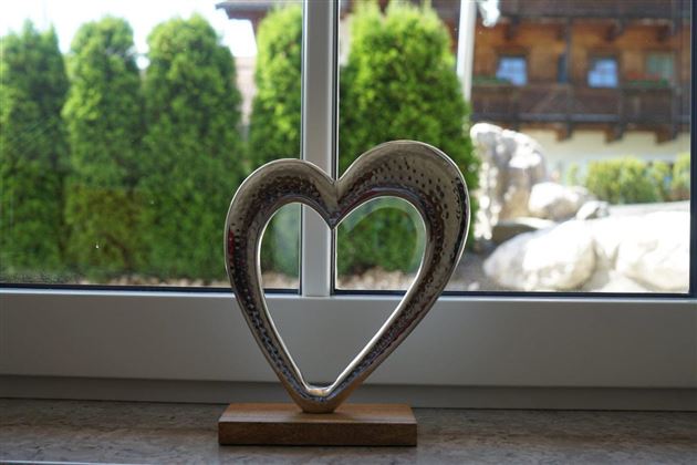 Een decoratieve hartschaal van zilver staat op een houten sokkel. Op de achtergrond zijn groene bomen en een deel van een huis te zien.