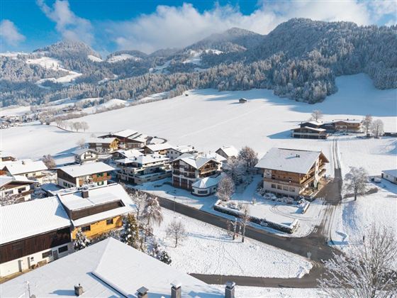 Eine malerische winterliche Landschaft mit verschneiten Häusern und sanften Hügeln im Hintergrund. Der klare blaue Himmel hebt die idyllische Szenerie hervor.