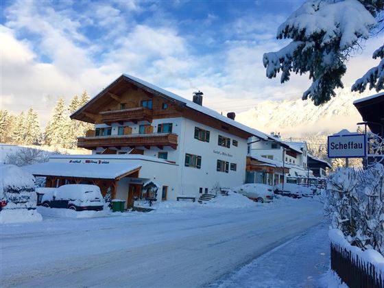 Eine verschneite Straße mit mehreren Häusern in einem Winterdorf. Die Umgebung ist malerisch mit Schnee bedeckten Bäumen und einem klaren Himmel.