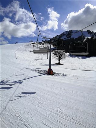 Eine Skiliftanlage auf einer verschneiten Piste unter einem klaren blauen Himmel. Im Hintergrund sind einige Berge und Wolken zu sehen.