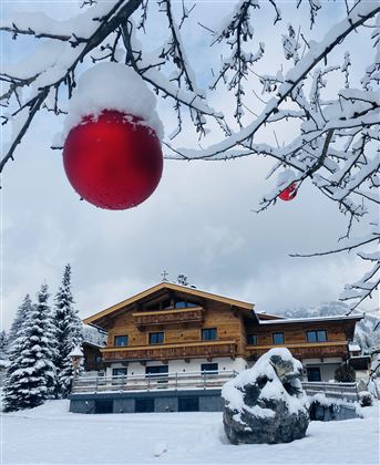 Ein schneebedecktes Landschaftsbild mit einem hübschen Holzhaus. Im Vordergrund hängt eine rote Weihnachtskugel an einem Baum.