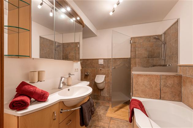 Ein modernes Badezimmer mit einer Dusche und einer Badewanne. Es gibt Holz- und Fliesenoberflächen sowie auffällige rote Handtücher.