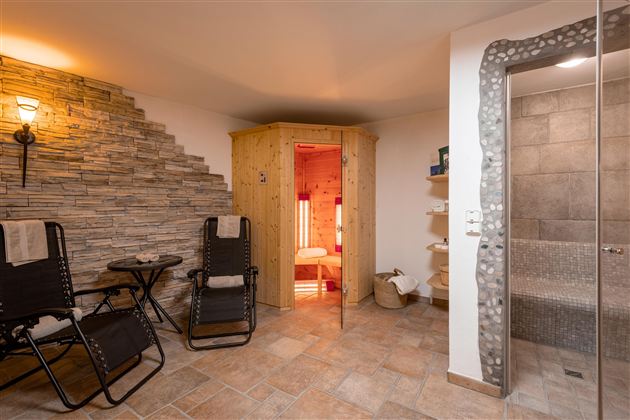 Ein gemütlicher Wellnessbereich mit einer Sauna, einem Dampfbad und stilvollen Liegestühlen. Die Wände sind mit Steinverkleidung dekoriert, und das Licht schafft eine entspannte Atmosphäre.