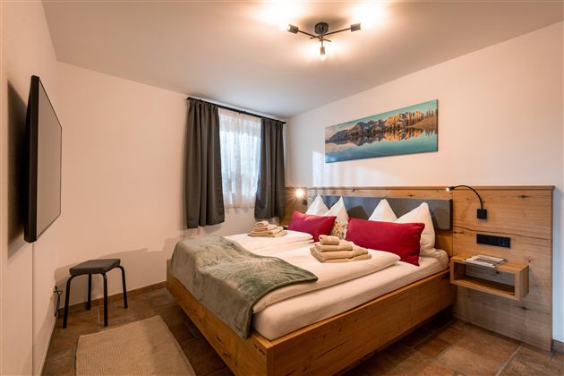 Ein modernes Schlafzimmer mit einem Doppelbett und holzfarbenen Möbeln. Der Raum ist hell mit einem großen Fenster und einem Fernseher an der Wand.