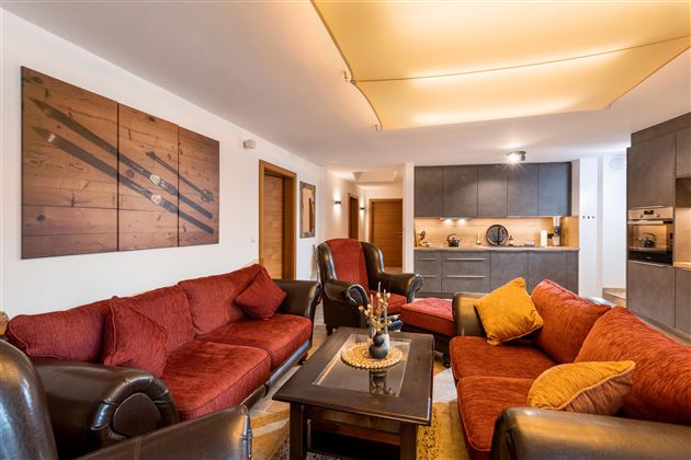 Ein modernes Wohnzimmer mit roten Sofas und einem eleganten Tisch. Die offene Küche ist im Hintergrund sichtbar und sorgt für eine gemütliche Atmosphäre.