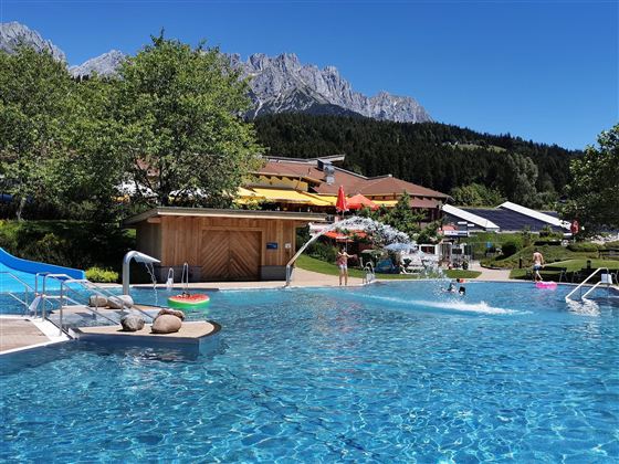 Ein schöner Pool mit klarem Wasser und einer sonnigen Umgebung. Im Hintergrund sind Berge und Grünflächen zu sehen.