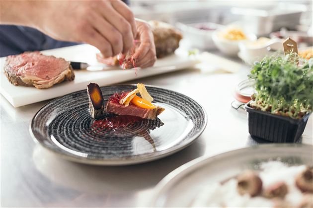 Een gevulde schotel met gefileerde vleespresentatie en kleurrijke garnering. Op de achtergrond zijn nog meer ingrediënten en keukengerei voor de bereiding te zien.