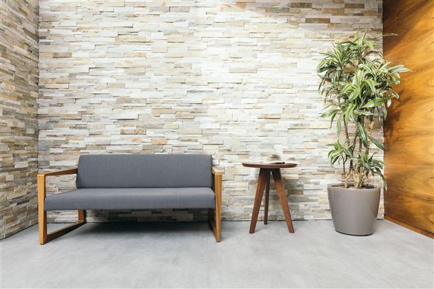 Een moderne wachtruimte met een grijze bank en een ronde houten tafel. Naast de bank staat een grote pot met een plant.