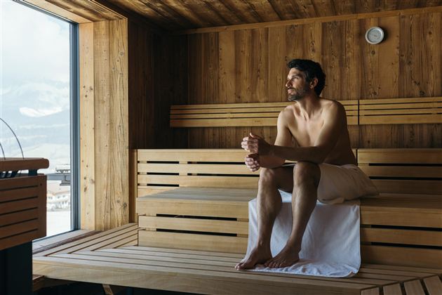 Een man zit in een houten sauna en kijkt uit het raam. De sfeer is ontspannen en gezellig.