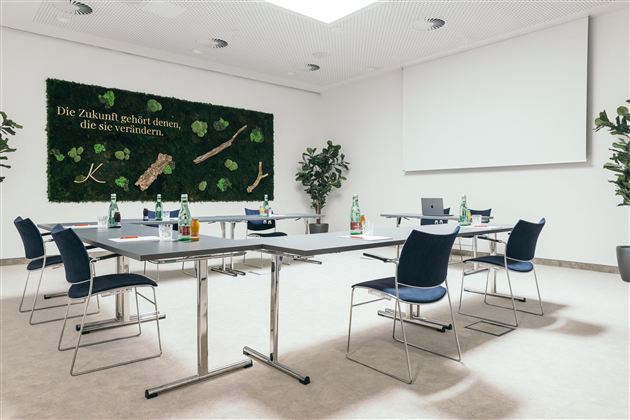 Een moderne vergaderruimte met tafels en stoelen. Aan de muur hangt een groene decoratieve oppervlakte met plantenmotieven en een inspirerende uitspraak.
