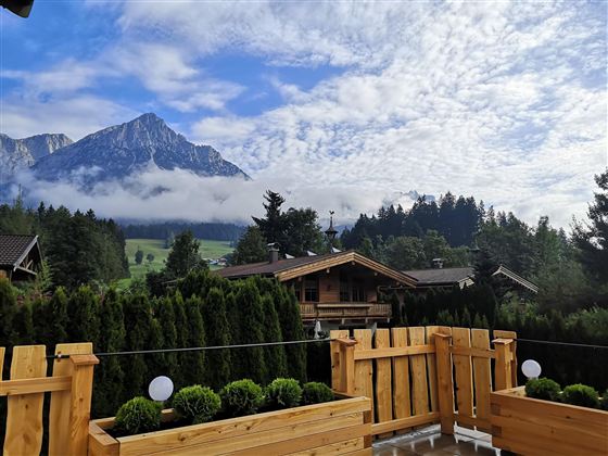 Eine malerische Berglandschaft mit hohen Gipfeln und bewölktem Himmel. Im Vordergrund eine Holzterrasse mit grünen Pflanzen und Blick auf angrenzende Häuser.