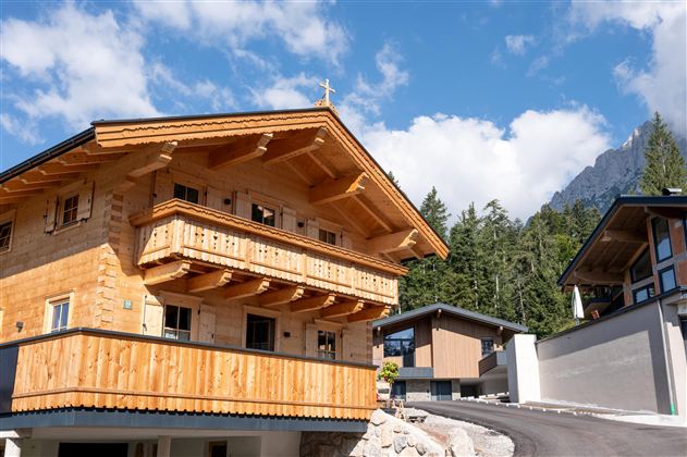 Een traditioneel houten huis in de bergen met een mooi balkon en het landschap van Kufstein op de achtergrond. De heldere lucht en de omringende bomen geven de scène een idylische sfeer.