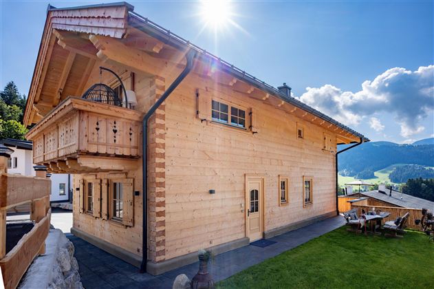 Een modern houten huis met een balkon en grote ramen. De omgeving is groen en heuvelachtig, met een stralend blauwe lucht.