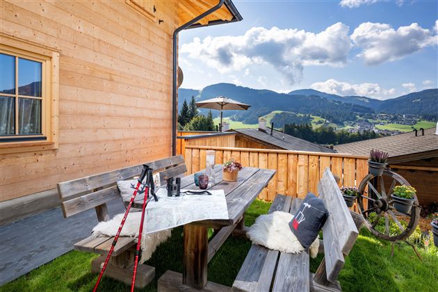 Een gezellige terras met een houten tafel en banken, omringd door een mooie berglandschap. Op de achtergrond zijn groene weiden en een blauwe lucht met wolken te zien.