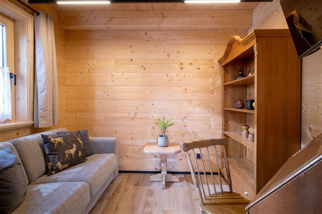 Een gezellige kamer met een grijze bank, een kleine tafel en een houten plank. Houten panelen aan de muren creëren een warme sfeer.