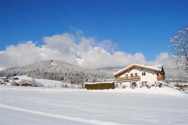 A snowy landscape with a charming house and majestic mountains in the background. The sky is clear and blue, creating a serene winter atmosphere.