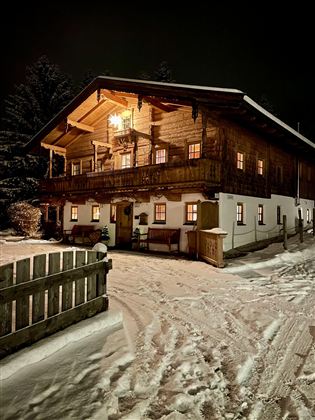 A cozy wooden house at night, illuminated by warm light. The path is covered with snow and the surroundings feel peaceful and inviting.