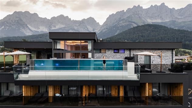 Een modern hotel met een infinity-pool op het dakterras. Op de achtergrond zijn indrukwekkende bergen te zien.