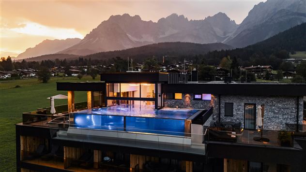 Een modern huis met een infinity-pool en uitzicht op de bergen. De scène wordt verlicht door een indrukwekkende zonsondergang.