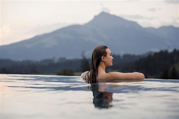 Een vrouw ontspant in een infinitypool met uitzicht op de bergen. De zachte avondstemming en het rustige landschap creëren een ontspannende sfeer.