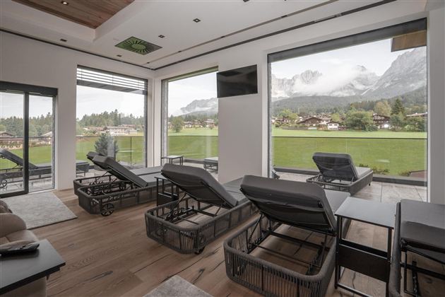 Een moderne ruimte met ligbedden en grote ramen die een groene landschap en bergen tonen. De ruimte voelt licht en uitnodigend aan.